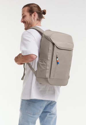 Uomo con capelli legati indietro che indossa una maglietta bianca e jeans, porta uno zaino beige grande con tasca frontale con cerniera e una linguetta colorata.