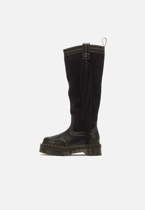 Stivale nero in pelle fino al ginocchio con suola spessa, cuciture decorative bianche e lunga frangia nera sul lato su sfondo semplice.