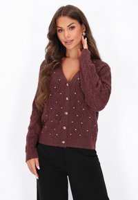 Femeie cu păr lung, castaniu, purtând un cardigan tricotat în nuanță de vișină, cu butoane și strasuri, asortat cu pantaloni negri, pozând cu o mână aproape de față.