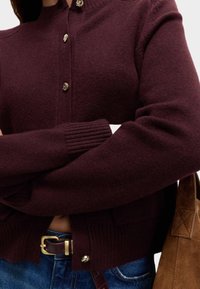 Burgundy stickad kofta med guldknappar, ribbade ärmslut och avslappnad passform; ihop med blå jeans och en handväska i brunt mocka.