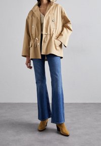 Chaqueta de algodón color beige con capucha, cintura ajustable con cordón, y dos bolsillos delanteros, combinada con jeans azules acampanados y botas de tacón marrones.