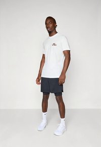 Weißes Baumwoll-T-Shirt mit kurzen Ärmeln, versehen mit einem roten "Just Do It"-Logo, kombiniert mit schwarzen Shorts und weißen Turnschuhen.