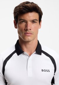 Weißes Polohemd mit schwarzen Akzenten, mit einem Kragen, drei Knöpfen und dem Wort "BOSS" auf der linken Seite gedruckt.