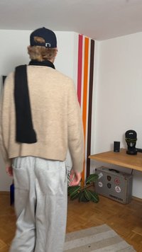 Beige strikket sweater med et sort tørklæde draperet over skulderen, grå joggingbukser. Baggrunden har farverige lodrette striber og stueplanter.