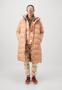 Pessoa em pé com um casaco puffer longo em pêssego, uma jaqueta de fleece estampada e calças, sapatos de caminhada vermelhos e óculos de sol desportivos refletivos, contra um fundo simples.