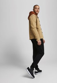 Giacca button-up beige con cappuccio marrone, pantaloni neri e sneakers nere. Tessuto morbido, vestibilità regolare, con dettaglio del cappuccio a contrasto e loghi minimali.