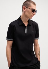 Camisa polo preta de mangas curtas, com um colarinho e punhos cinzentos, e um logótipo discreto no peito. Textura suave e design ajustado.