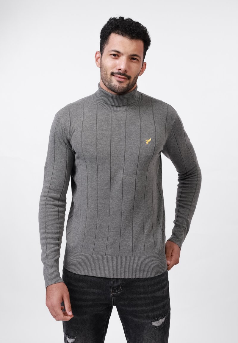Mann mit kurzen, lockigen Haaren, der einen grauen, gerippten Rollkragenpullover und schwarze, zerrissene Jeans trägt, steht vor einem weißen Hintergrund.