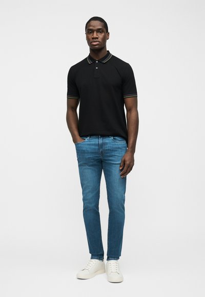 Μαύρο polo με κοντό μανίκι και γιακά με ρίγες σε χρώματα του ουράνιου τόξου, μπλε στενά τζιν και λευκά αθλητικά παπούτσια που φοριέται από έναν άνδρα που στέκεται με το ένα χέρι στην τσέπη.