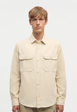 Młody mężczyzna o jasnobrązowych włosach, ubrany w beżową koszulę zapinaną na guziki z dwiema kieszeniami na piersi, stoi na tle jednolitego jasnego tła.
