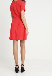 Robe rouge à pois blancs avec manches courtes, taille ajustée, jupe évasée et texture lisse. Assortie à des sandales noires à talons hauts.