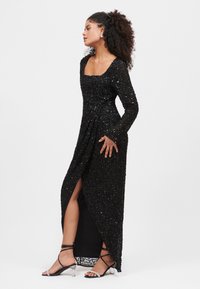 Abito da sera nero in paillettes con maniche lunghe, scollo quadrato, spacco laterale e tessuto testurizzato. Abbinato a sandali con tacco trasparente.
