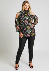 Blusa nera floreale con maniche trasparenti a spalla fredda; fiori vibranti rosa e gialli; abbinata a leggings neri lucidi e scarpe col tacco nere con cinturini.