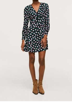 Frau trägt ein schwarzes, langärmeliges Kleid mit V-Ausschnitt und Blumenmuster, kombiniert mit braunen Wildleder-Stiefeletten, steht vor einem einfarbigen Hintergrund.