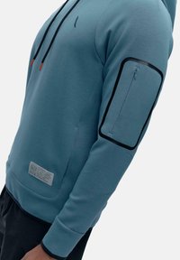 Modra kapuca z žametom z črnim žepom na zgornjem delu roke, dodatnim sprednjim žepom in belo oznako blagovne znamke na dnu.