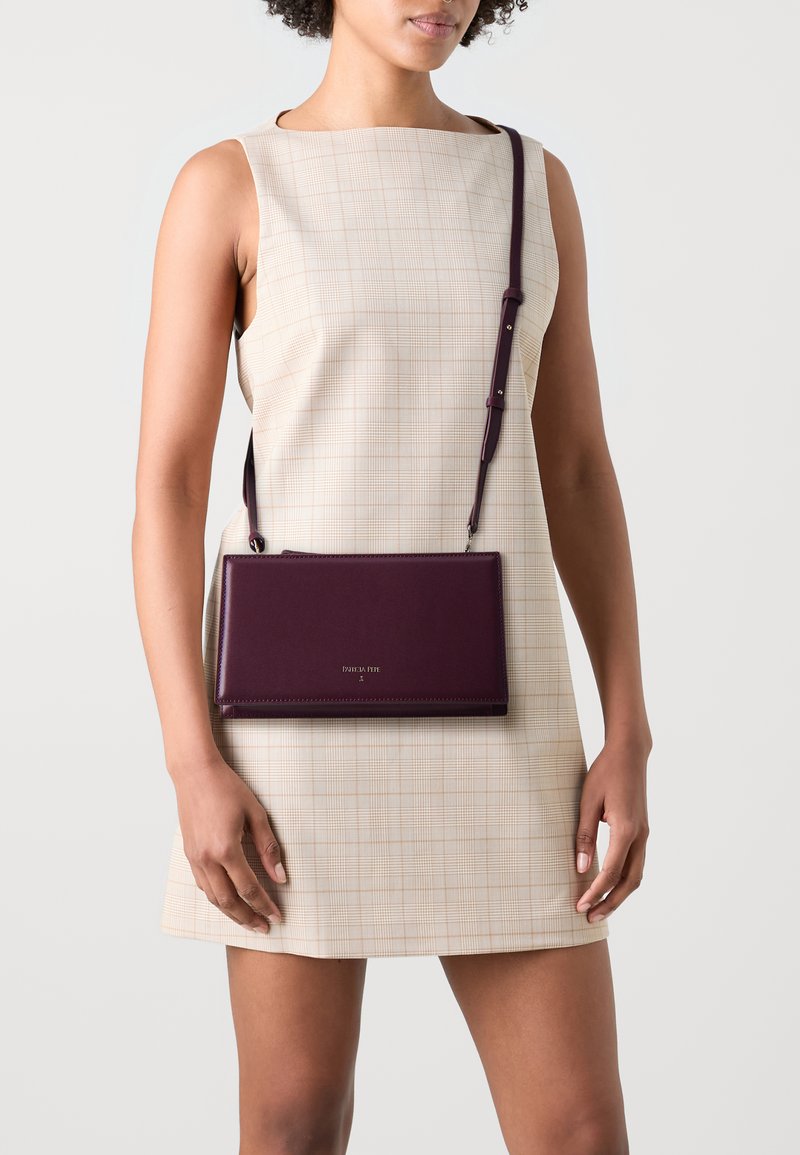 Taisnstūra formas purpursarkans ādas crossbody soma ar regulējamu siksnu, kurai ir logo detaļa. Modelis valkā beige krāsas svītrainu bez piedurknēm kleitu.