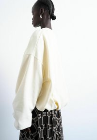 Femme aux cheveux tressés portant un pull crème surdimensionné et une jupe noire à motifs, debout de profil devant un fond blanc uni.