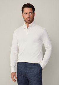Weißes Langarm-Polohemd mit einer sanften Textur, ausgestattet mit einem spitzen Kragen und einer Knopfleiste mit drei Knöpfen. Kombiniert mit dunkelblauen gemusterten Hosen.
