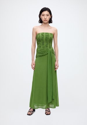 Mujer con el pelo corto y negro que lleva un vestido verde sin tirantes con fruncidos y una cinta drapeada, de pie delante de un fondo blanco liso.