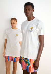 T-shirts brancos de algodão apresentam um gráfico de sol e onda no peito; calções de banho coloridos com padrões de limões, riscas e designs abstratos.