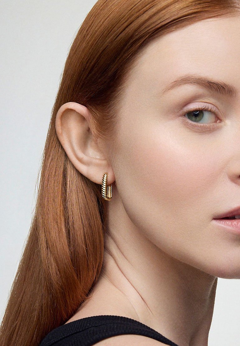 Złote kolczyki w kształcie kół z teksturowanym wzorem, charakteryzujące się gładkim wykończeniem i lekko zakrzywionym kształtem, noszone przez modelkę z prostymi, rdzawymi włosami.