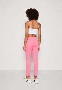 Pantalon rose ajusté avec une texture lisse et une coupe slim, doté de poches arrière et d'une étiquette beige à la taille, associé à des sandales blanches à talons blocs.