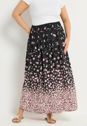 Frau trägt einen schwarzen Maxirock mit pink-weißem Blumenmuster, weißes kurzärmliges Oberteil und beige Sandalen mit offener Spitze.