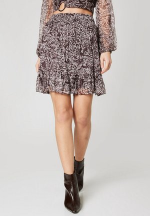 Mujer con minifalda estampada con volantes, mangas largas transparentes y botas negras hasta el tobillo, de pie sobre un fondo claro y liso.