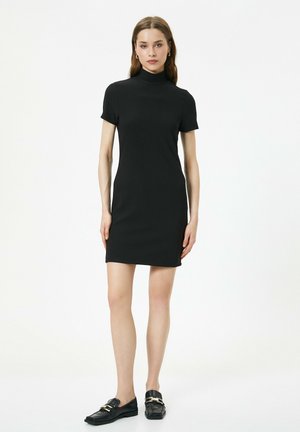 Schwarzes Kurzarmkleid mit hohem Kragen, gefertigt aus glattem Stoff. Kombiniert mit dunklen Loafers mit goldenen Akzenten.