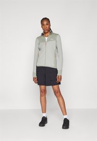 Giacca zip-up verde chiaro con texture a costine e dettagli neri, abbinata a pantaloni cargo neri e sneakers nere. Design semplice e funzionale.