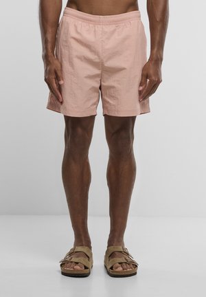 Man draagt lichtroze korte broek en beige sandalen, staat tegen een effen lichtgrijze achtergrond, toont het onderlichaam en de benen.