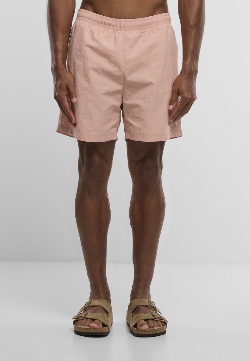 Homme portant un short rose clair et des sandales beige, debout devant un fond uni gris clair, montrant le bas du torse et les jambes.
