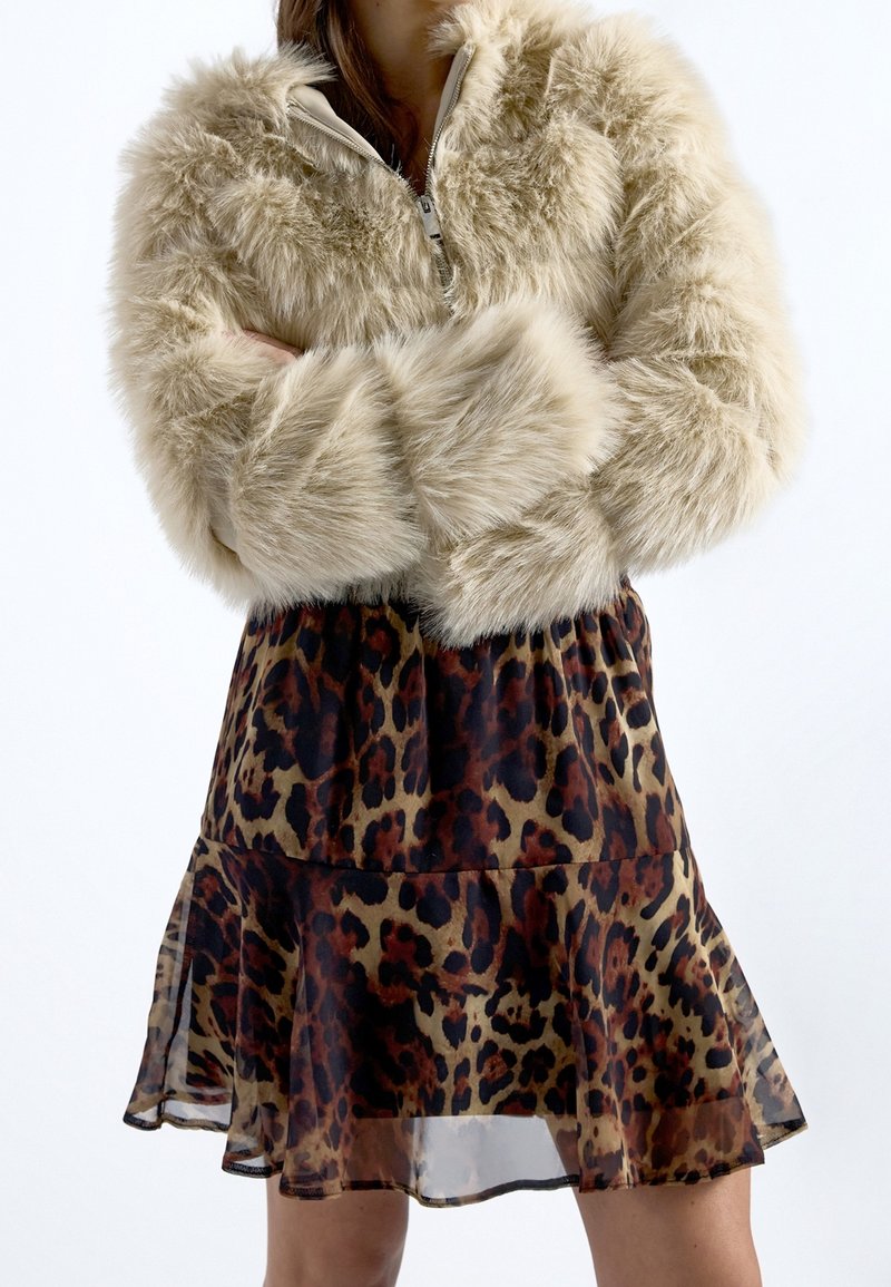 Personne portant une veste en fausse fourrure beige zippée, associée à une jupe courte à imprimé léopard, bras croisés sur la poitrine.