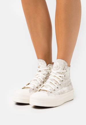 Sneakers wsuwane w jasnobeżowym materiale z kwiecistym wzorem, z białymi sznurowadłami, zaokrąglonym noskiem i teksturowaną gumową podeszwą.