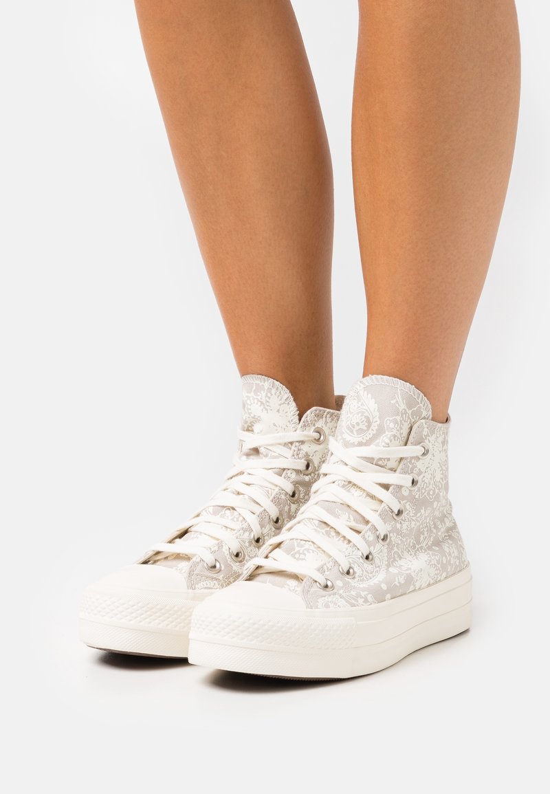 Sneakers wsuwane w jasnobeżowym materiale z kwiecistym wzorem, z białymi sznurowadłami, zaokrąglonym noskiem i teksturowaną gumową podeszwą.