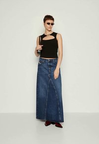 Mulher vestindo uma blusa preta sem mangas com recorte, uma saia longa de denim azul, botas vermelhas, uma pequena mala preta e óculos de sol finos pretos.
