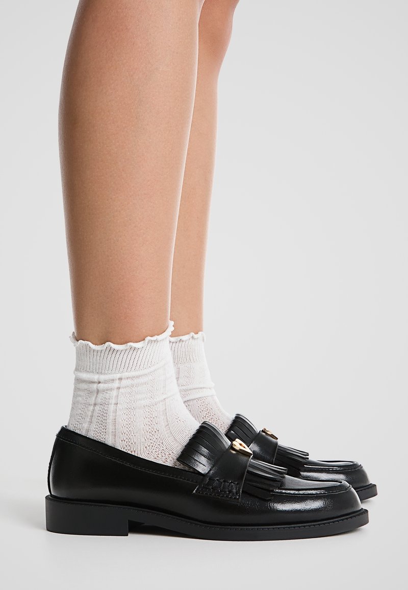 Jambes portant des chaussettes blanches texturées à la cheville associées à des mocassins en cuir noir à pampilles sur un fond neutre.