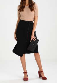 Jupe crayon noire avec fermeture éclair devant, associée à un haut beige côtelé. Les accessoires incluent une pochette noire avec des détails floraux et des escarpins rouges.