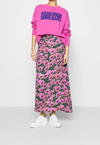 Pull fuchsia avec texte bleu marine, associé à une jupe longue imprimée florale avec des fleurs roses sur un fond vert foncé, et des baskets blanches.