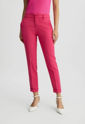 Mujer vestida con pantalones tobilleros de color rosa brillante a medida con dobladillos doblados y tacones blancos de punta puntiaguda con correa en el tobillo, posando contra un fondo liso.