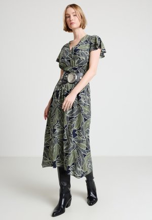 Donna che indossa un vestito scuro con motivi di foglie verdi, cintura larga con borchie e stivali neri al ginocchio, in piedi davanti a uno sfondo bianco uniforme.