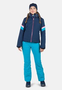 Blaue Skijacke mit kontrastierenden pinken und türkisfarbenen Streifen, kombiniert mit türkisfarbenen Hosen. Verfügt über einen Reißverschluss und einen Stehkragen.