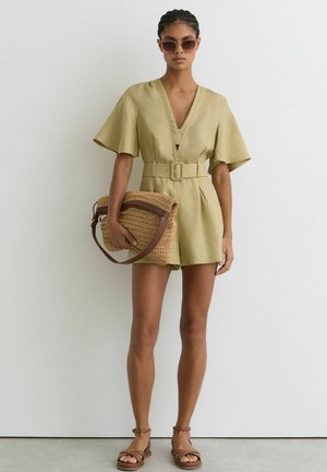 Kvinde iført beige romper med bælte, brune sandaler, solbriller og holder en stor flettet håndtaske mod en ensfarvet hvid baggrund.