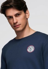 Marineblå langærmet skjorte lavet af bomuld med rund halsudskæring. Har en cirkulær logo-patch med røde og hvide accenter på brystet.