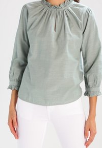 Light green cotton blouse with a gathered neckline, keyhole opening, and three-quarter sleeves featuring ruffled cuffs.