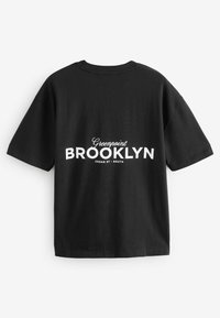 Melna kokvilnas T-kreklu ar noapaļu kakla izgriezumu, īsām piedurknēm un baltu uzrakstu "Greenpoint BROOKLYN CEDAR ST - SOUTH" uz muguras.