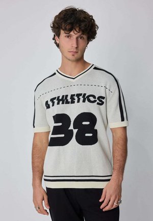 Jeune homme aux cheveux bouclés portant un pull en tricot blanc avec des rayures noires, le texte "ATHLETICS" et le numéro "38" sur le devant, debout devant un fond gris.