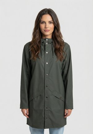 Donna con capelli castano lunghi che indossa un impermeabile verde scuro con cappuccio, bottoni automatici e tasche frontali, in piedi davanti a uno sfondo uniforme.