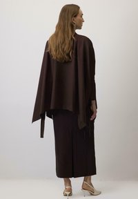 Poncho di stoffa marrone con design drappeggiato, indossato sopra una gonna lunga coordinata. L'outfit presenta una texture sottile e accenti minimalisti.
