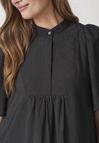 Mustast pullover-tüüpi pluus ümmarguse kraega, millel on nööpidega kinnitusriba, volditud kujundus ja lühikesed läikivad varrukad. Valmistatud siledast kangast.